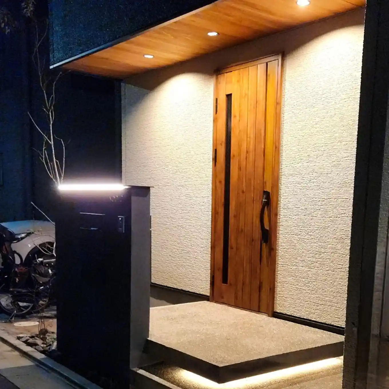 木製ドアと間接照明が特徴的な夜の住宅エントランス。シンプルで高級感のある外観。